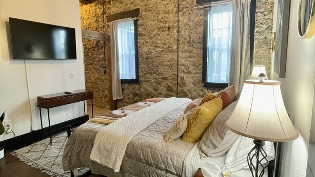 A bedroom with a bed, a desk with a lamp, a TV, and stone walls. - Historic 1 bed 1 bath in 1920's Chevy (Mountain Home, AR, 72653)