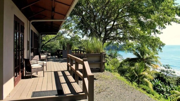 Una terraza de madera con mobiliario exterior, con vistas a una zona costera con árboles y al mar.