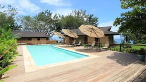 Una terraza de madera con piscina, sillones de descanso y sombrillas.
