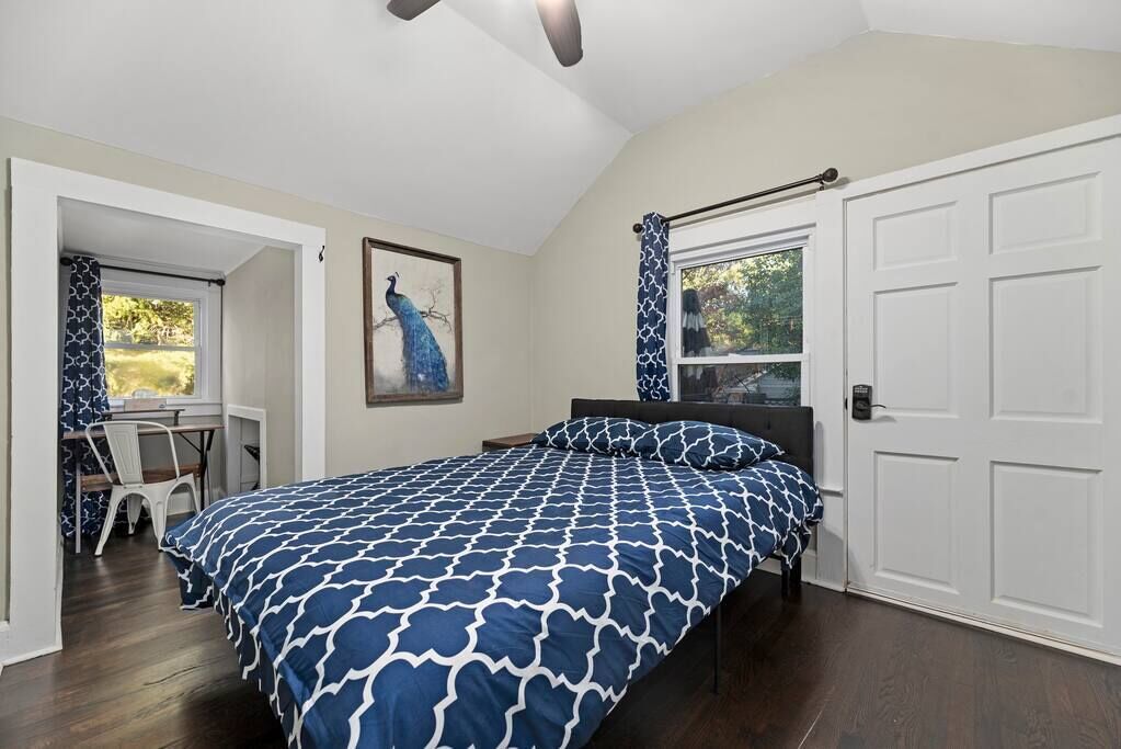 Une chambre à coucher avec un lit, un bureau et une chaise. Il y a une fenêtre donnant sur des arbres et une porte.