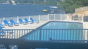 En swimmingpool med blå solstolar, ett vitt staket och utsikt över havet och avlägsna byggnader.