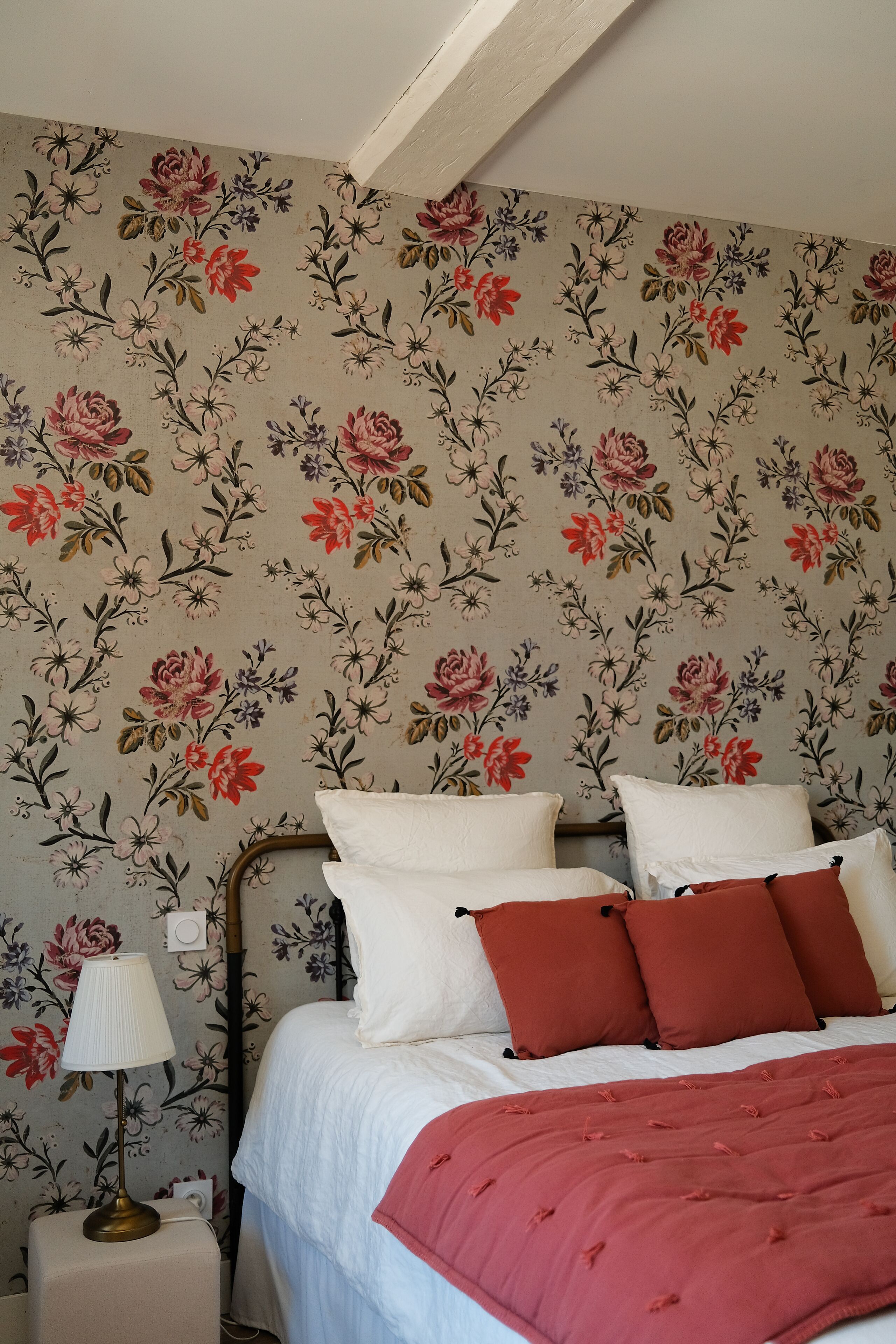 A bedroom with floral wallpaper, a bed with white and red bedding, and a bedside lamp.