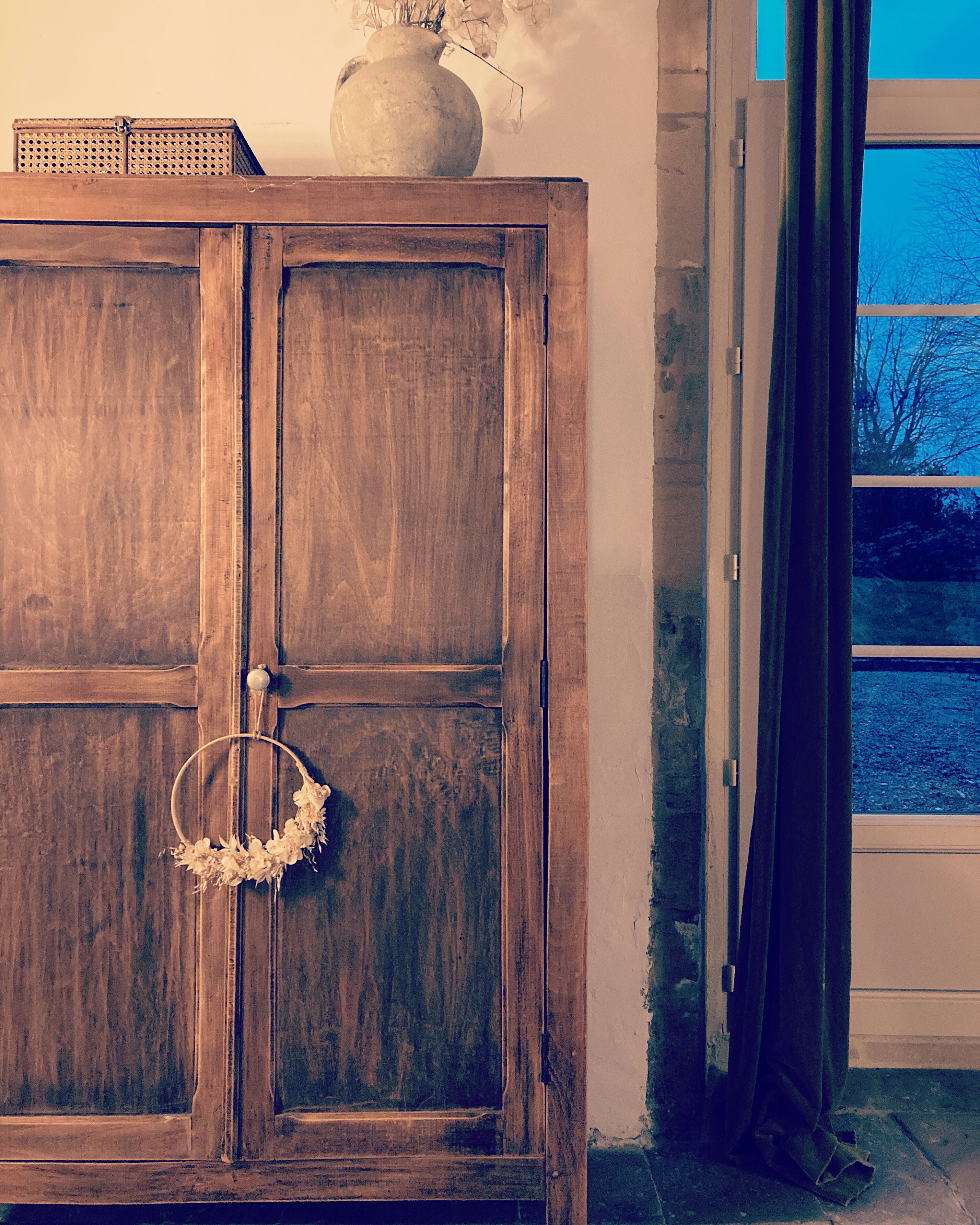 Un mueble de madera con un adorno de guirnalda, un jarrón con flores secas y una ventana con cortinas.