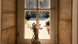 Une pièce dotée d’une grande fenêtre, d’un vase contenant des fleurs séchées posé sur une table en bois, et offrant une vue sur un bâtiment à l’extérieur.