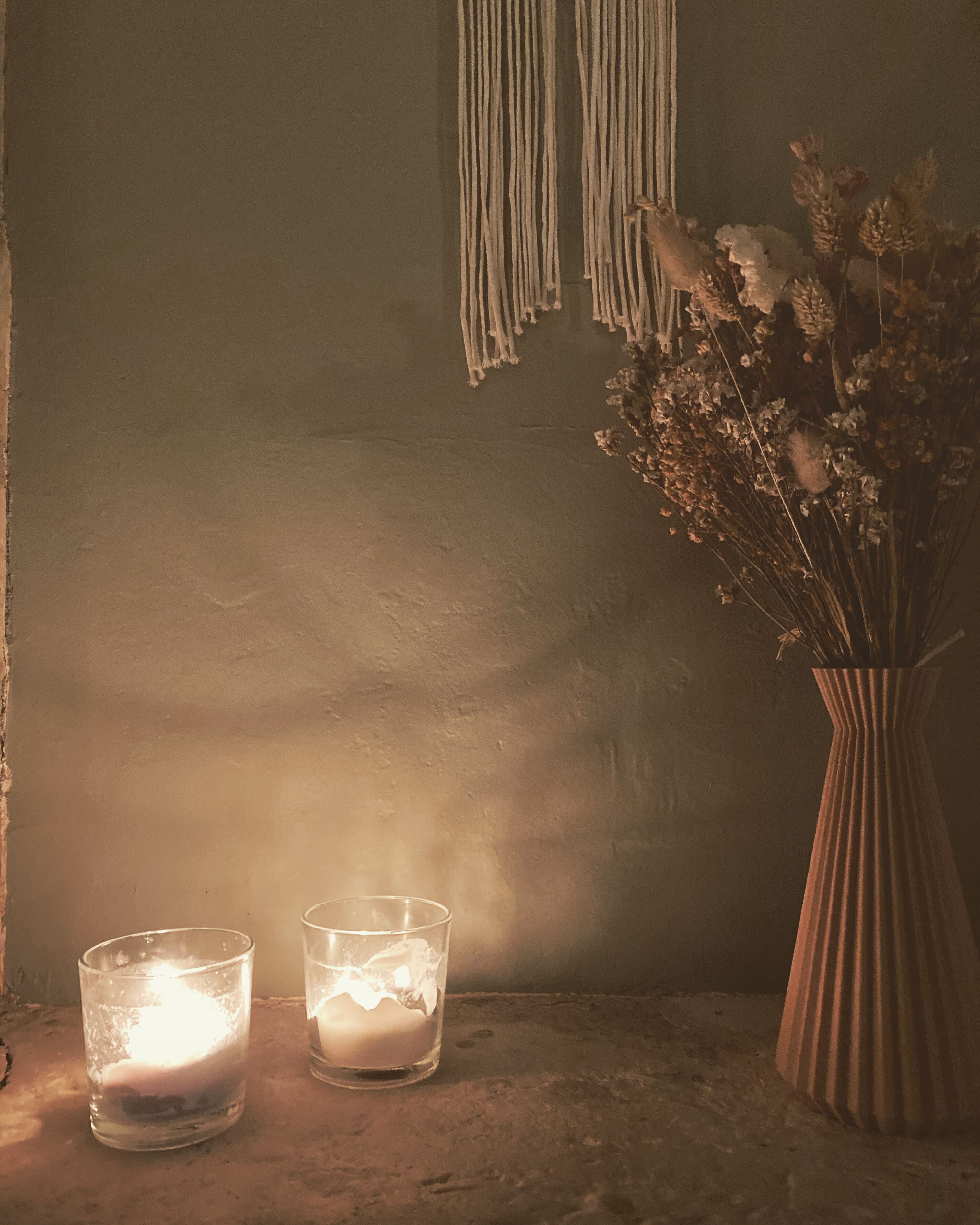 A candlelit table with two candles and a vase of dried flowers against a textured wall.