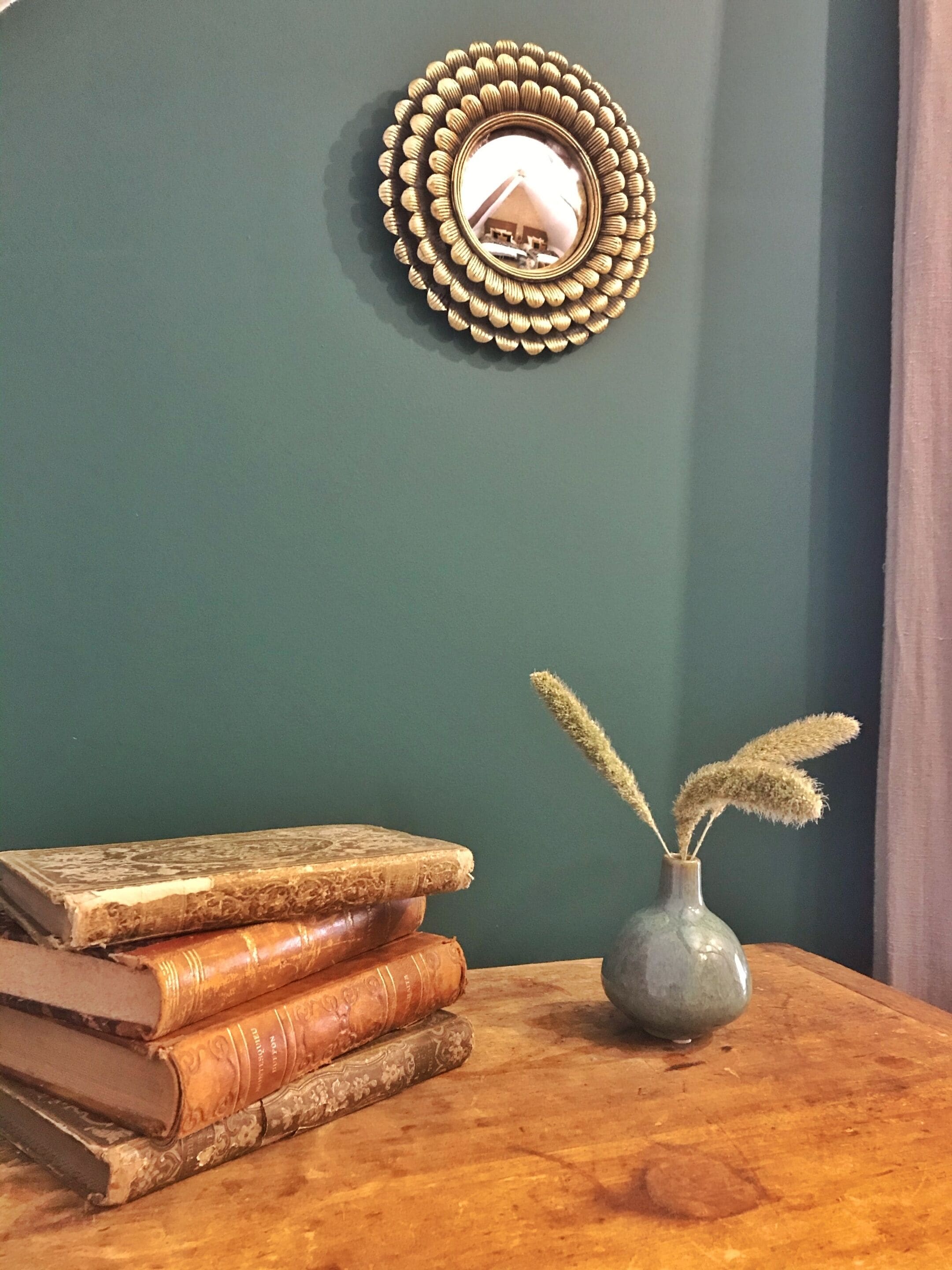 Une pile de livres sur une table en bois, un vase contenant des plantes séchées et un miroir décoratif sur un mur vert.