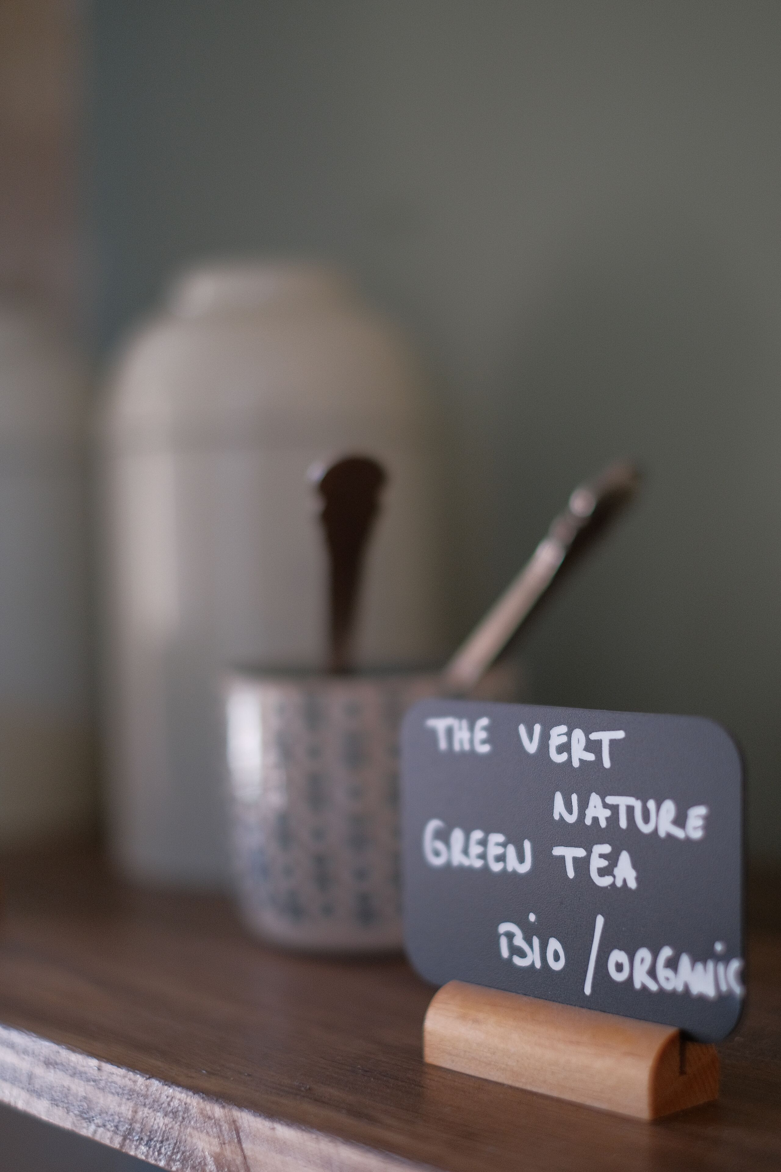 Een houten oppervlak met een bordje waarop staat 'THE VERT NATURE GREEN TEA BIO / ORGANIC' en een lepel in een keramieken pot.
