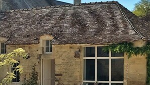 Un bâtiment en pierre avec un toit en tuiles, une grande fenêtre et une cour en gravier aménagée avec du mobilier d’extérieur.