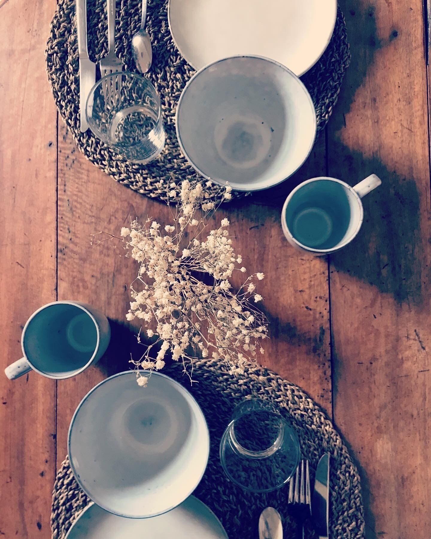 Una mesa de comedor dispuesta con platos, tazones, tazas y cubiertos sobre un camino de mesa tejido.