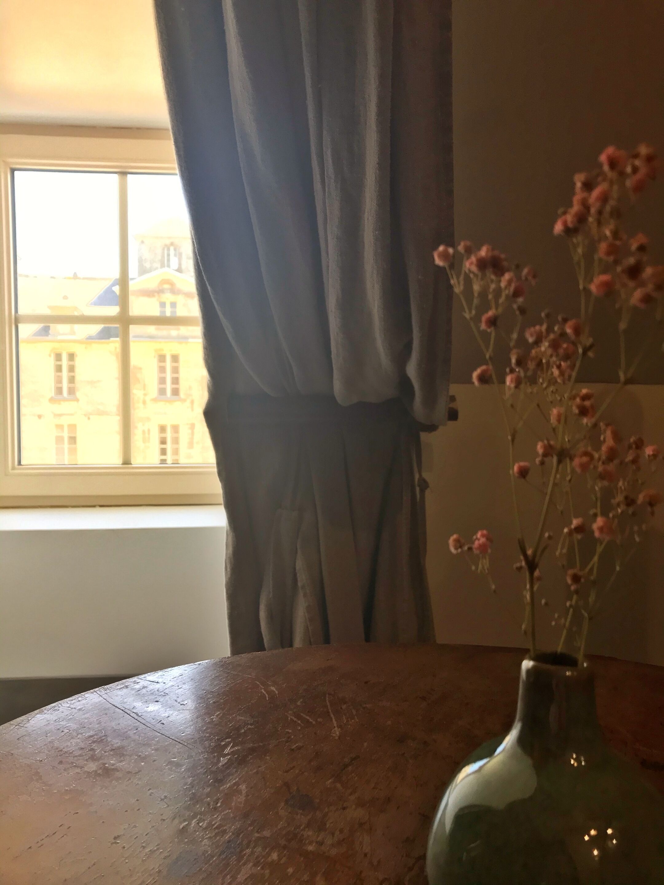 Una habitación con ventana, cortina y un jarrón con flores sobre una mesa.