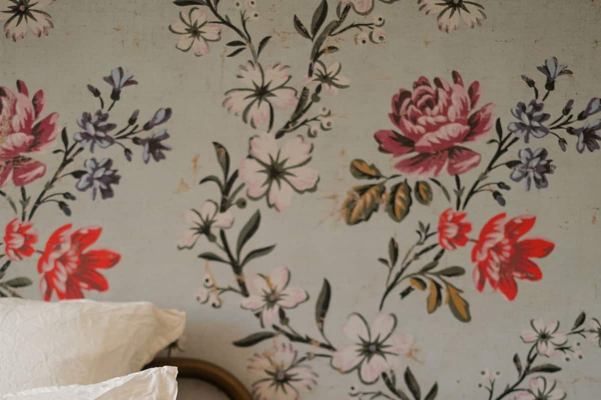 Une chambre à coucher avec un papier peint à motifs floraux, un lit avec une literie blanche et rouge, une lampe en laiton et une table de chevet blanche.