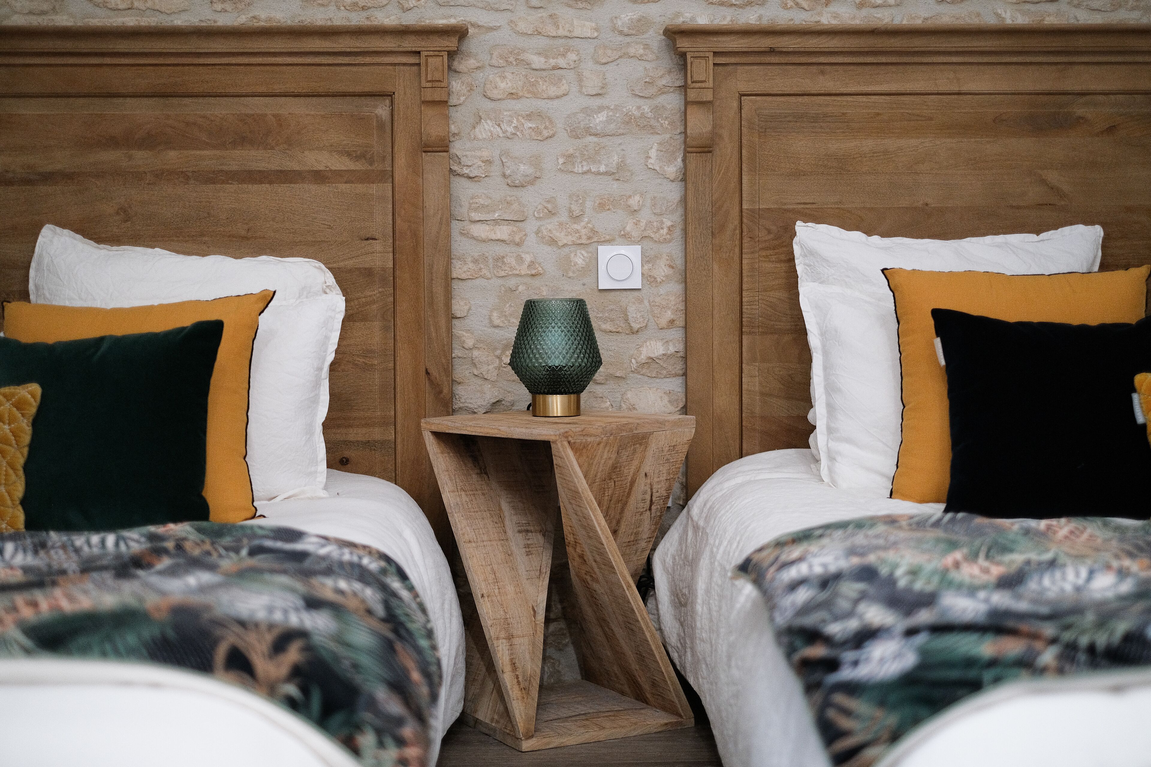 Une chambre à coucher avec deux lits, chacun agrémenté d’oreillers blancs et colorés, et une petite table de chevet en bois avec une lampe verte placée entre les deux.