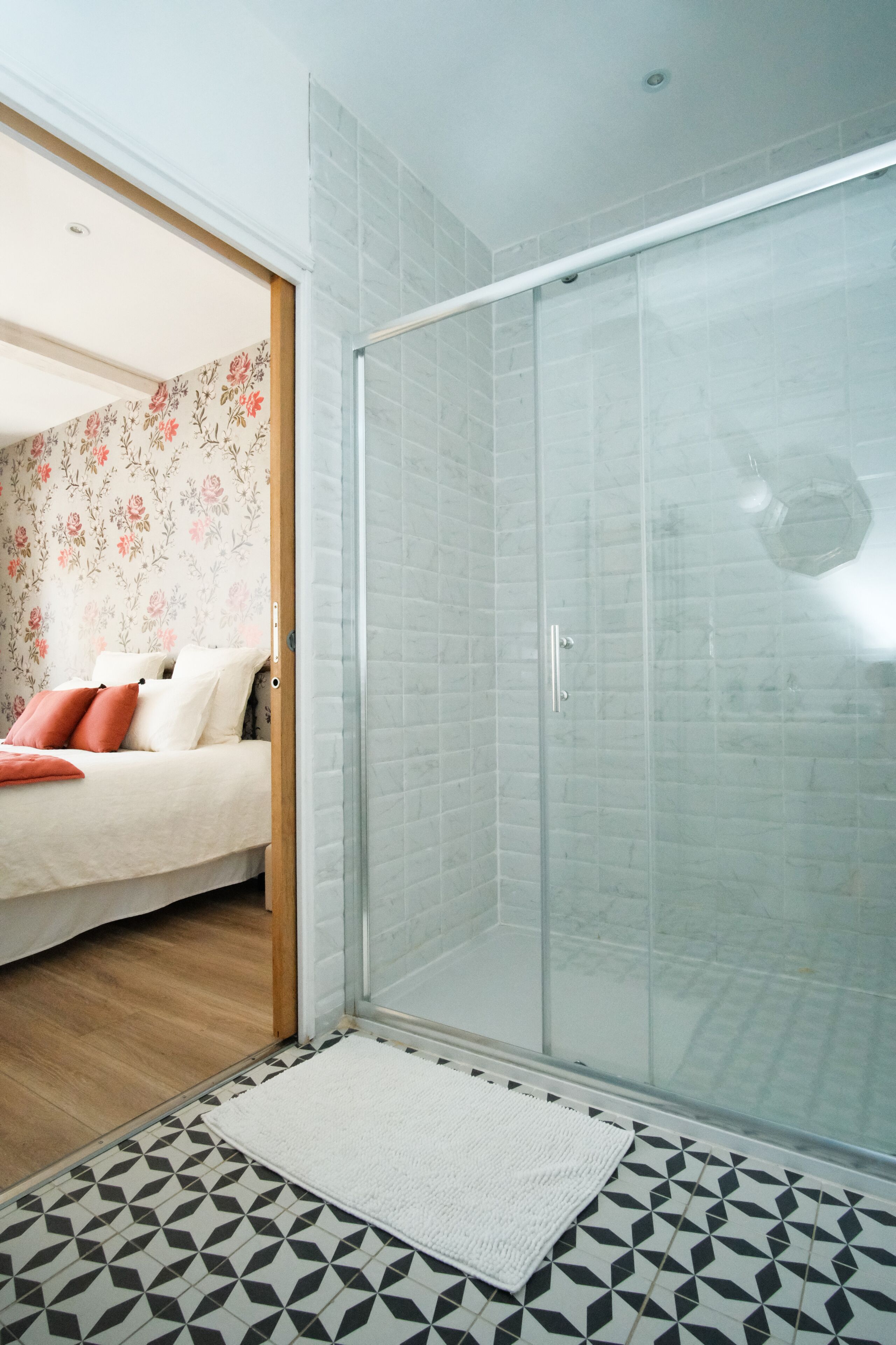 A modern bathroom with a glass shower enclosure, a patterned floor, and a bed with floral wallpaper in the adjacent room.