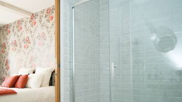 A modern bathroom with a glass shower enclosure, a patterned floor, and a bed with floral wallpaper in the adjacent room.