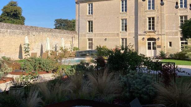 Un bâtiment en pierre doté d’une grande fenêtre et d’un balcon, entouré d’un jardin avec des allées de gravier et des plantes.
