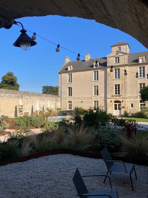 Un bâtiment en pierre doté d’une grande fenêtre et d’un balcon, entouré d’un jardin avec des allées de gravier et des plantes.