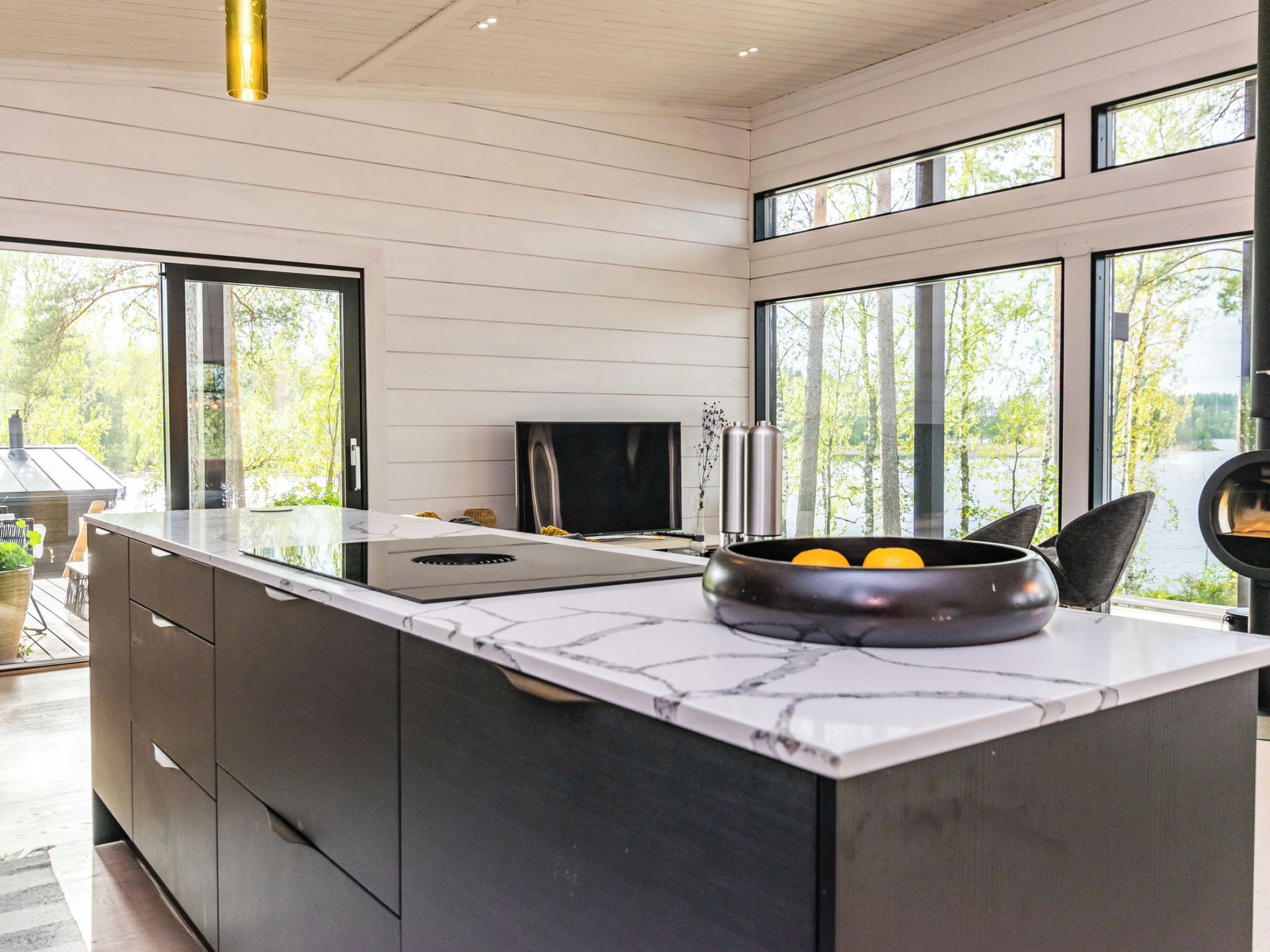 A modern kitchen with a large island, a flat-screen TV, and a view of a lake through large windows.