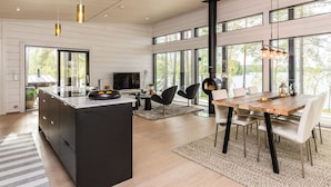 Eine geräumige, moderne Küche mit einer großen Kochinsel, einem Essbereich mit Holztisch und -stühlen sowie großen Fenstern mit Blick ins Freie.