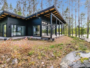 Moderni, kaksikerroksinen mökki, jossa on suuret ikkunat, puinen terassi ja metsäinen tausta.