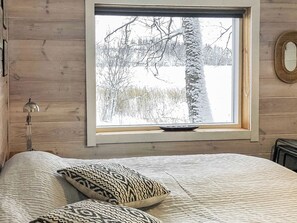 Makuuhuone, jossa on suuri ikkuna, puuseinät, kuviollisilla tyynyillä varustettu sänky ja yöpöydällä oleva lamppu.