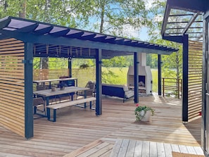 Un patio extérieur couvert avec une terrasse en bois, une table avec des chaises, un banc et un espace barbecue.