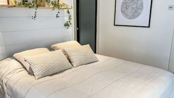 A bed with a quilted white duvet and two pillows, a wooden shelf with plants, a door, and a framed abstract artwork on the wall.