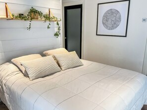Ein Bett mit weißer, gesteppter Bettdecke und zwei Kissen, ein Holzregal mit Pflanzen, eine Tür und ein gerahmtes abstraktes Kunstwerk an der Wand.