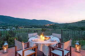 A round table with chairs set for dining, candles, and a bottle, overlooking a mountainous landscape.