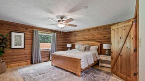 A bedroom with a wooden log wall, a bed with a headboard, a ceiling fan, a nightstand with a lamp, and a large wooden door. - Welcome to Holly Hill – peaceful lakefront living (Denver)