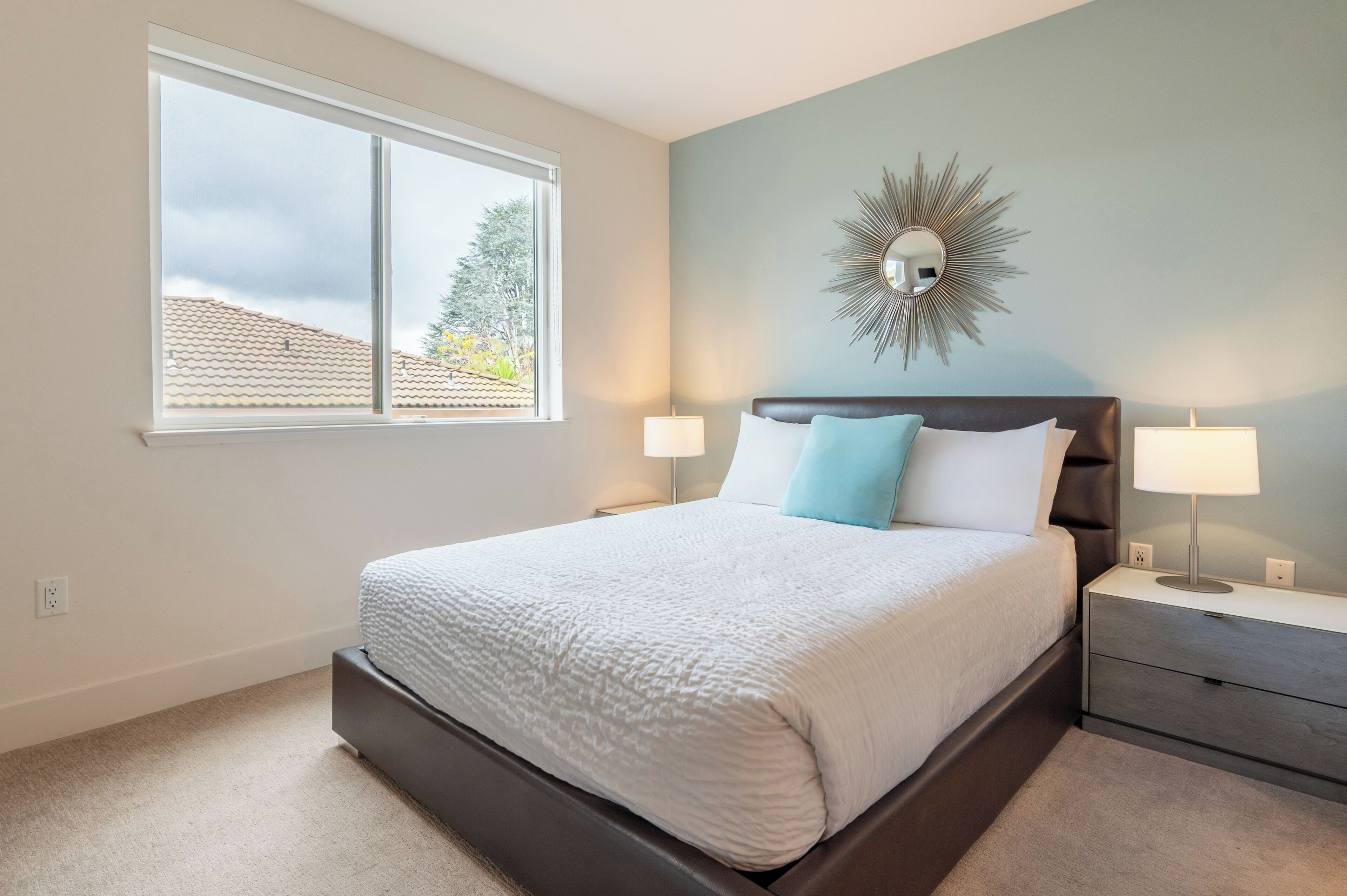 A bedroom with a large window, a bed with white and blue bedding, a dark headboard, two bedside tables with lamps, and a wall-mounted mirror.