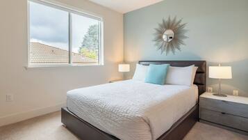 A bedroom with a large window, a bed with white and blue bedding, a dark headboard, two bedside tables with lamps, and a wall-mounted mirror.