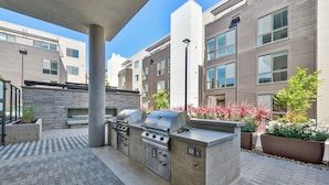 An outdoor patio with a built-in grill and seating area, surrounded by modern buildings. - Luxury One Bedroom with Full-Service Amenities (Mountain View)