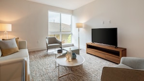 A modern living room with a sofa, armchair, coffee table, TV, and floor lamp. - Luxury One Bedroom with Full-Service Amenities (Mountain View)
