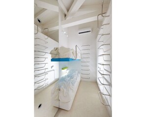 Una habitación estrecha y blanca con una cama individual superpuesta, barandillas de metal y un pequeño equipo de aire acondicionado.