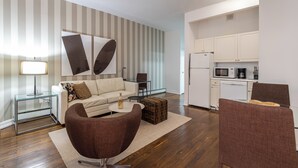 Un salon moderne avec un canapé, un fauteuil et une table basse, une cuisine avec des meubles blancs et des appareils électroménagers, et un mur à rayures.