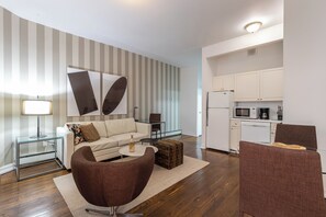 Un salon moderne avec un canapé, un fauteuil et une table basse, une cuisine avec des meubles blancs et des appareils électroménagers, et un mur à rayures.