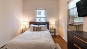 Une chambre à coucher avec un lit, deux tables de chevet avec des lampes, un téléviseur fixé au mur et une fenêtre avec des stores.