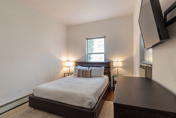 Un dormitorio con cama, lámparas de noche, una ventana y un televisor de pantalla plana montado en la pared.