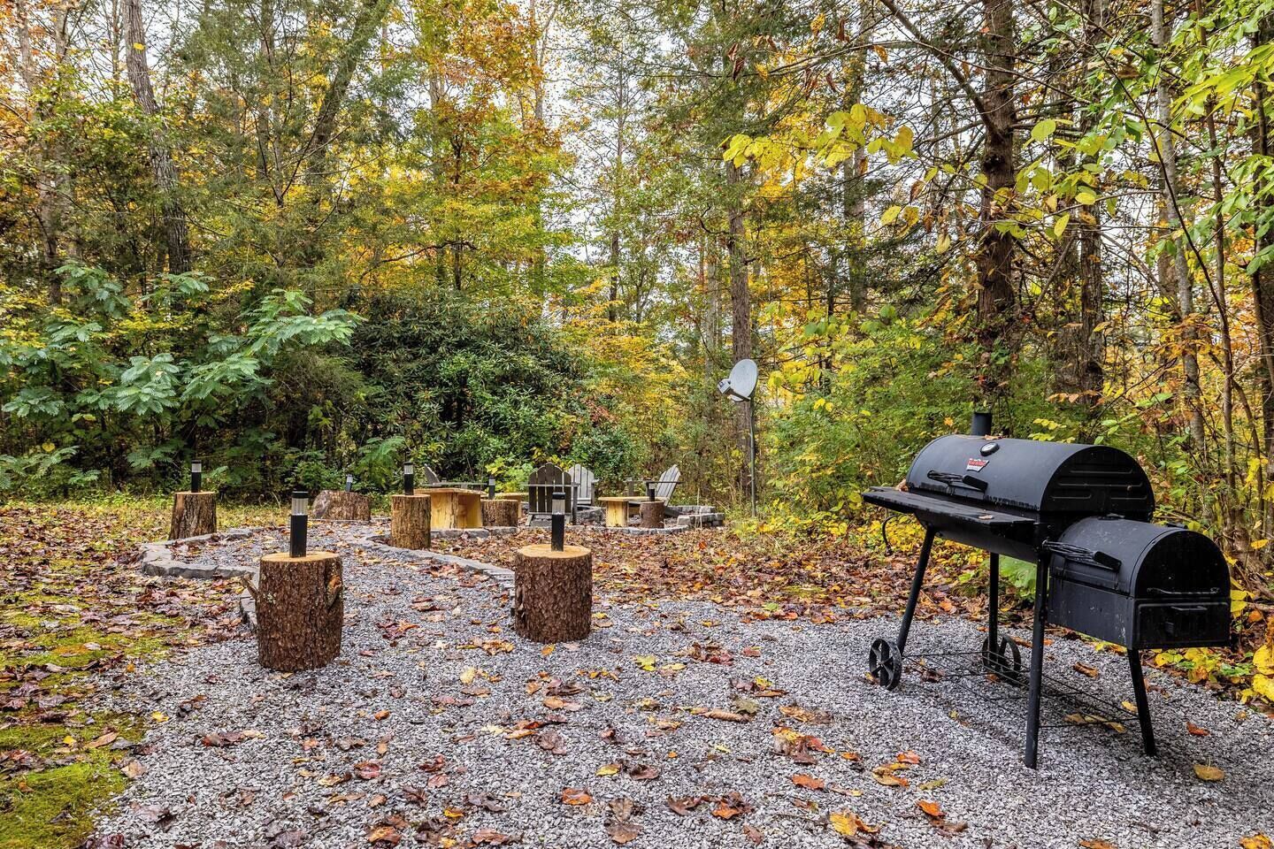 A rustic outdoor area with a grill, wooden posts, and a gravel path surrounded by trees.