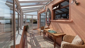 A sunlit room with a glass ceiling, a wicker sofa, a wooden table, and a view of the sea.