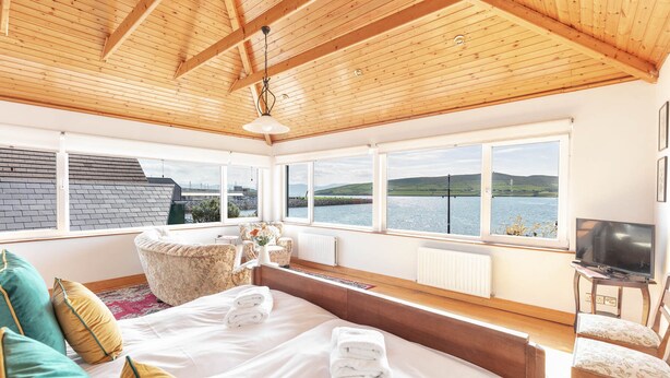 A bedroom with a large bed, a sofa, a chair, and a TV. There are windows with a view of the water and hills.