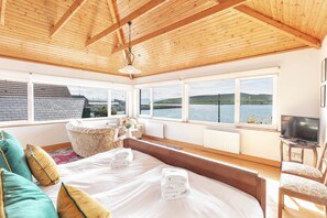 A bedroom with a large bed, a sofa, a chair, and a TV. There are windows with a view of the water and hills.