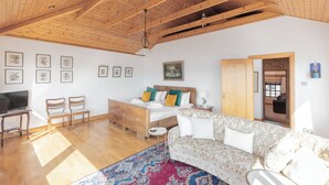A spacious living area with a high wooden ceiling, a sofa, a bed with colorful pillows, a TV, and framed artwork on the walls.