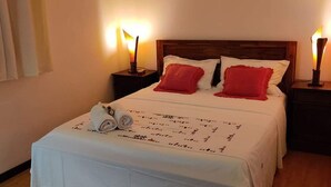 A neatly made bed with white linens and red pillows, two bedside lamps, an air conditioning unit on the wall, and a window with curtains.