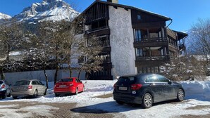 Un immeuble à plusieurs étages doté d’un balcon, avec des voitures garées et un sol recouvert de neige.