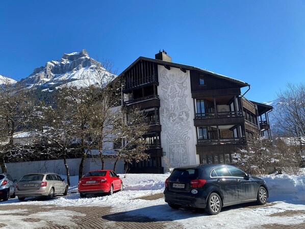 A multi-story building with a balcony, parked cars, and snow-covered ground.