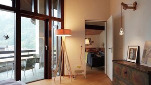 Une chambre spacieuse avec une grande fenêtre, un lampadaire, un tapis à motifs et une commode en bois sur laquelle est posé un cadre photo.