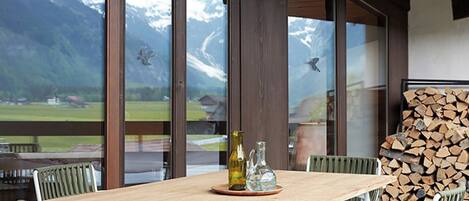 A wooden table with four chairs, a bottle, and a pitcher on a coaster, set against a backdrop of a mountain view through large windows.