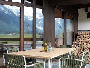 Une table en bois, quatre chaises, une bouteille et un pichet posés sur un dessous de verre, le tout devant une vue sur les montagnes, visible à travers de grandes fenêtres.