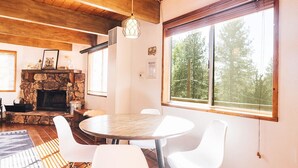 Una habitación acogedora con chimenea de piedra, vigas de madera en el techo, una mesa redonda con sillas blancas y un ventanal con vistas a árboles.